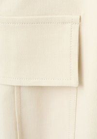 Primer plano de un bolsillo de solapa de tela beige con costuras visibles sobre material tejido texturizado.