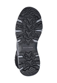 Suela de zapatilla Skechers negra con textura de goma, presenta profundos surcos y diseños angulares para tracción. Logotipo visible en el lateral.