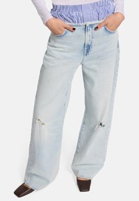 Himmelblaue, weit geschnittene Denim-Jeans mit Fronttaschen, die unregelmäßige Details in der Nähe der Knie aufweisen. Kombiniert mit einem gestreiften Stoffbund.