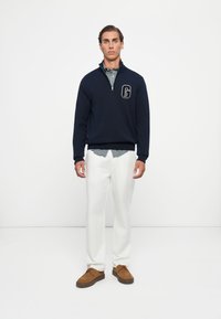 Navy zip-neck sweater with a 'G' logo, layered over a green checkered shirt, paired with white pants and brown suede shoes.