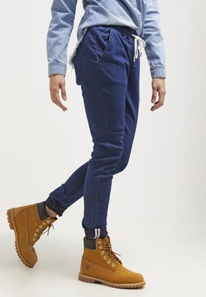 Person trägt marineblaue Jogginghose, hellblaues Jeanshemd und hellbraune Schnür-Arbeitsschuhe, steht mit einem leicht gehobenen Fuß.