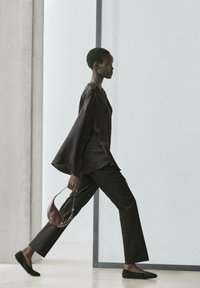 Modella con top nero a maniche lunghe e ampie, pantaloni neri e scarpe basse nere, che tiene una borsa marrone mentre cammina.