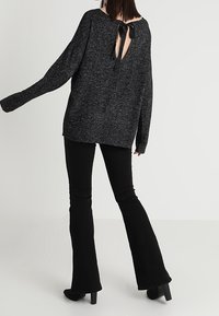 Černý pletený svetr s dlouhými rukávy a volným střihem, který má velký zadní výstřih svázaný černou stuhou. Spárován s černými zvonovými kalhotami.