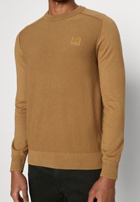 Brun stickad tröja med ett texturerat mönster, rund halsringning och ribbade muddar. Har en liten präglad logotyp på bröstet.