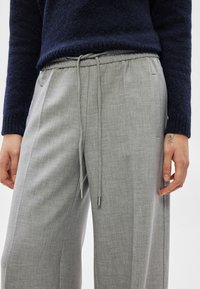 Sieviete valkā gaiši pelēkas bikses ar aukliņu un tumši zilu pūkainu džemperi, ar mazu tetovējumu redzamu uz kreisās rokas.