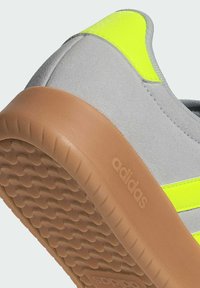 Gray suede sneaker with neon yellow accents, rubber brown outsole, textured grip, and visible branding on the heel.