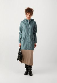 Vodoodporna turkizno zelena jakna s kapuco, srebrnimi sponkami, dvema sprednjima žepoma, nošena preko bež krila in črnih škornjev do kolen. V roki drži črno torbico.