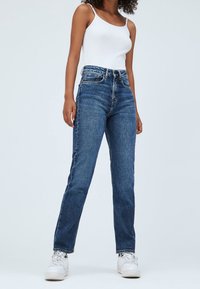 Vaqueros de tiro alto y pierna recta en azul con textura de denim, que cuentan con un diseño de cinco bolsillos y herrajes de botón metálico, combinados con una camiseta sin mangas blanca.