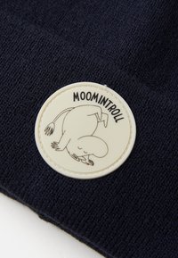 Căciulă tricotată navy cu o patch rotund, de culoare crem, ce prezintă un design de personaj de desene animate și textul "MOOMINTROLL". Textură moale și fit confortabil.