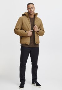 Giacca imbottita marrone con cappuccio, chiusura frontale con zip e dettagli neri. Indossata sopra un maglione marrone texturizzato, abbinata a pantaloni neri e sneakers.