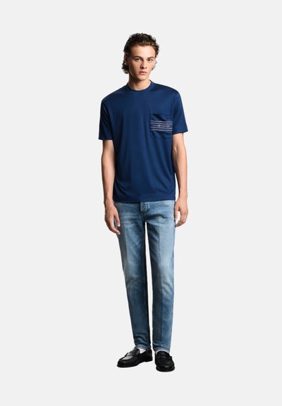 T-shirt blu navy a maniche corte con taschino anteriore caratterizzato da un motivo a righe, abbinato a jeans azzurri e scarpe nere.