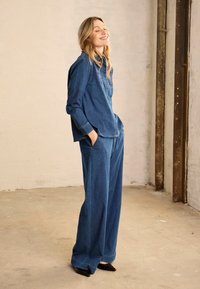 Femme portant une chemise en denim et un pantalon en denim à jambes larges, debout les mains dans les poches, souriante, devant un mur beige et une poutre métallique industrielle.