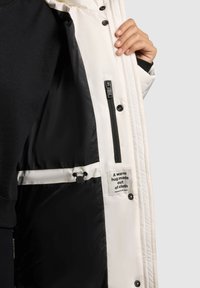 Jachetă izolată albă cu interior negru, buzunar lateral cu fermoar, detalii cu șnur și un etichetă care spune „O îmbrățișare caldă făcută din material.”