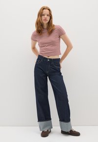 Jonge vrouw met rood haar, gekleed in een rood-wit gestreepte crop top, wijde, omgeslagen donkere jeans en bruine sneakers, staande tegen een witte muur.