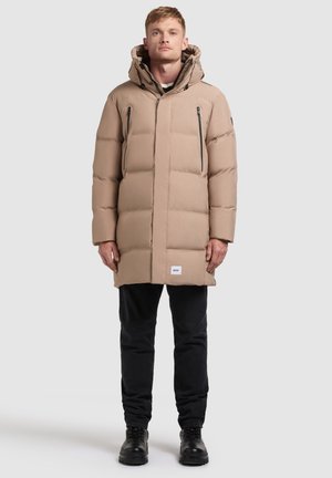Hombre vestido con un abrigo acolchado beige con capucha, pantalones negros y botas negras, de pie contra un fondo claro y liso.