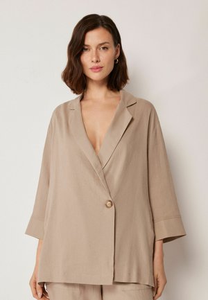Femme aux cheveux bruns mi-longs portant un blazer beige ample à bouton unique et manches trois-quarts, sur un fond clair.
