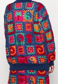 Cardigan all'uncinetto colorato con design a patchwork e motivi vivaci. Base blu, polsini e orlo a coste. Texture variegate e dettagli intricati.