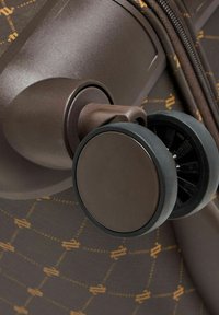 Gros plan sur des roues doubles pivotantes fixées à un tissu de valise marron à motifs avec une fermeture éclair visible en arrière-plan.