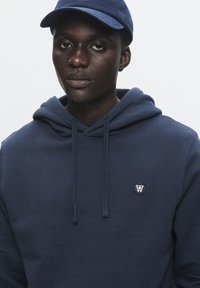 Marineblå hættetrøje med snore og broderet logo på venstre bryst; båret med en matchende marineblå baseballhat. Glat tekstur, afslappet pasform.