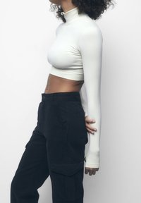 Personne portant un top court à manches longues blanc et un pantalon cargo noir, se tenant de côté contre un fond blanc uni.