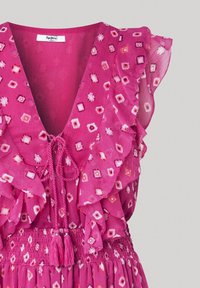 Różowa sukienka z głębokim dekoltem w kształcie litery V, falbanianymi rękawami i marszczonym pasem. Materiał ma kolorowy geometryczny wzór i ozdobne frędzle.