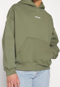 Homme portant un sweat à capuche vert olive avec une poche avant et un petit texte blanc « WRSTBHVR » au-dessus de la poitrine, associé à un jean bleu clair.
