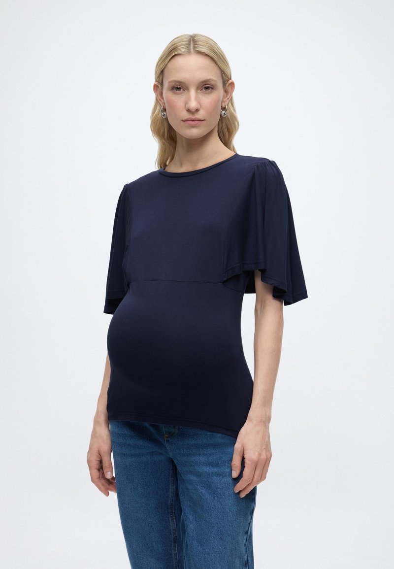 Έγκυος γυναίκα με ξανθά μαλλιά φοράει μπλε μπλούζα με μανίκια με κύματα και μπλε τζιν, στέκεται μπροστά σε απλό λευκό φόντο.