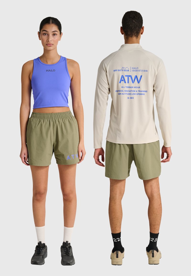 Eine Frau in einem blauen bauchfreien Top und olivgrünen Shorts blickt nach vorne; ein Mann trägt ein beige langärmeliges Hemd und olivgrüne Shorts und blickt nach hinten, beide tragen Turnschuhe.