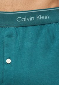 Κοντινό σε ζώνη βερμούδας Calvin Klein σε τυρκουάζ χρώμα με λευκό κουμπί και τμήμα γυμνής επιδερμίδας από πάνω.