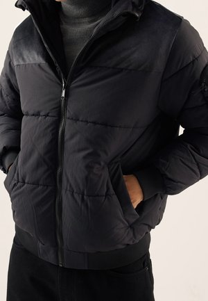 Personne portant une veste matelassée noire par-dessus un pull à col roulé gris foncé, les mains dans les poches de la veste.