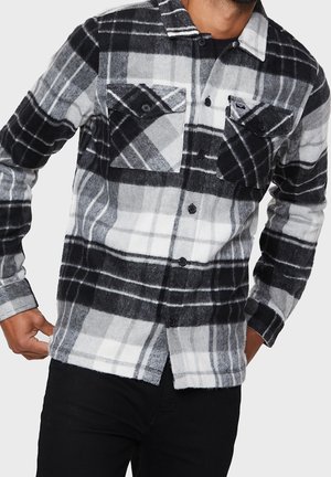 Camisa de franela a cuadros en negro y gris con dos bolsillos en el pecho, cierre de botones y acentos en blanco. Textura suave con un diseño de corte relajado.