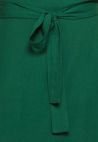 Tissu vert avec un détail de nœud papillon noué, montrant une texture lisse et des extrémités pendantes verticales sur le vêtement.