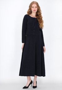 Tengerészek két részes szett, amely egy cropped kardigánt és hosszú szoknyát tartalmaz, mindkettő texturált szövetből készült, fekete magassarkú cipővel párosítva.