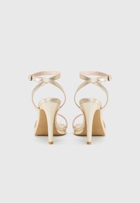Goldene High Heel Sandalen mit gekreuzten metallischen Riemen, einem Schnallenverschluss am Knöchel und einem spitzen Zehendesign mit einer strukturierten Oberfläche.