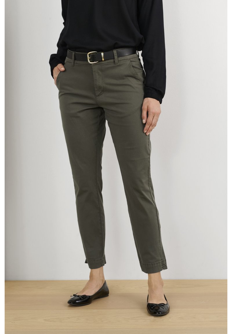 Femme portant un pantalon slim vert olive avec une ceinture noire, un top noir à manches longues et des ballerines noires, se tenant sur un sol en bois.