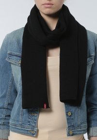 Bufanda de punto negra colocada alrededor del cuello, con una textura suave y una etiqueta de marca roja, llevada sobre una blusa beige y una chaqueta de mezclilla.