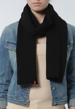 Bufanda de punto negra colocada alrededor del cuello, con una textura suave y una etiqueta de marca roja, llevada sobre una blusa beige y una chaqueta de mezclilla.