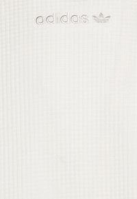 Valkoinen ribbikangas, jossa on brodeerattu vaaleanharmaa Adidas-logo ja kolmonen symboli yläkeskellä.