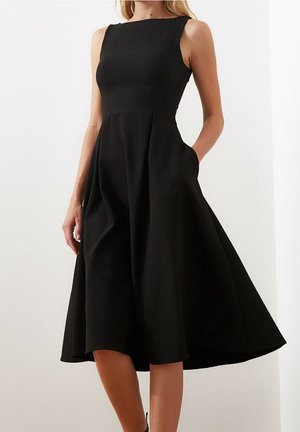 Ärmelloses schwarzes Midikleid mit figurbetontem Oberteil, ausgestelltem Rock und Seitentaschen, getragen von einer stehenden Frau vor einfachem Hintergrund.