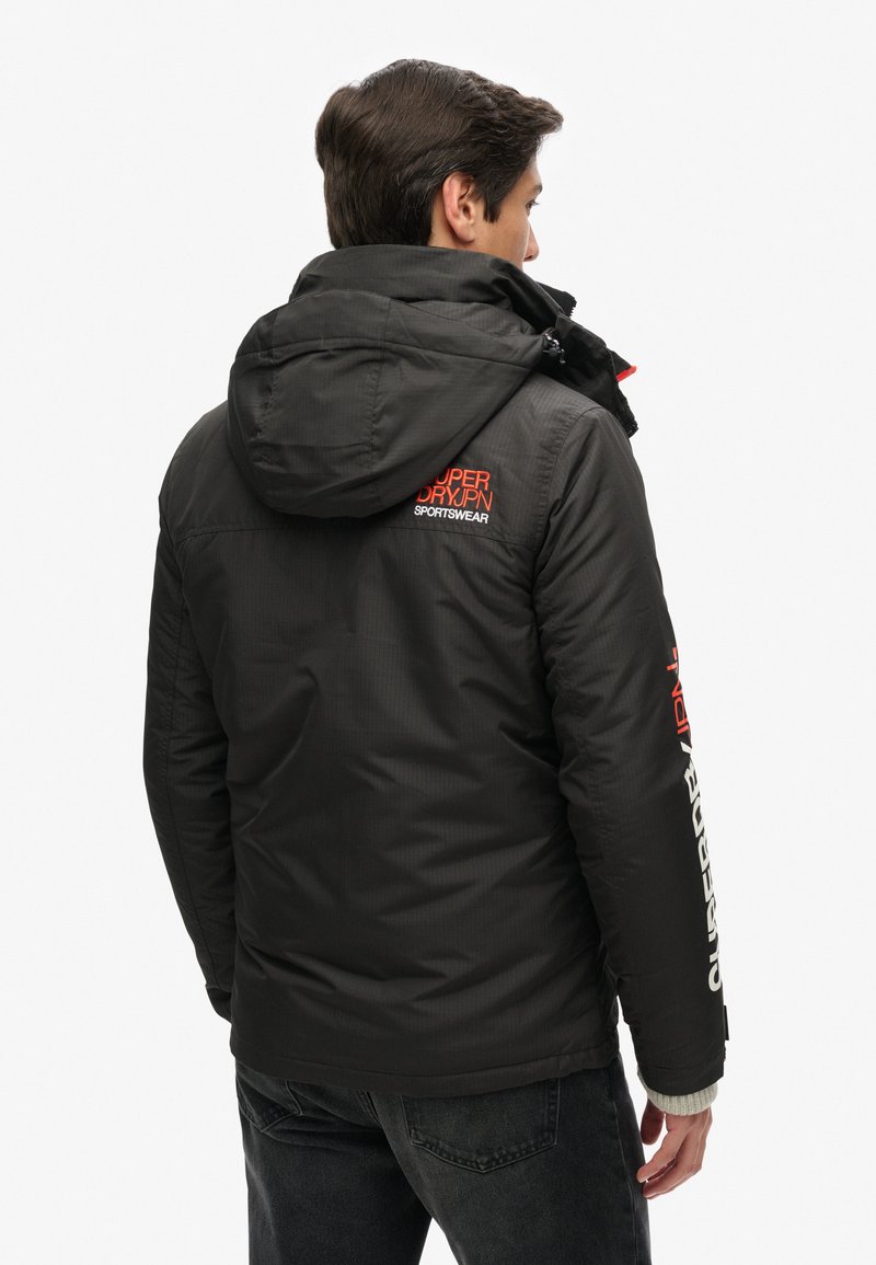 Chaqueta aislante negra con capucha, que presenta una marca roja y blanca en la parte posterior y en la manga, tejido texturizado y diseño resistente al viento.