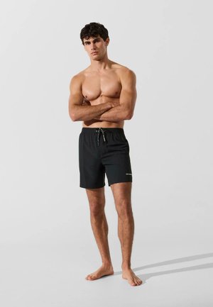 Ing nélküli férfi áll mezítláb, karját keresztbe téve, fekete térdig érő fürdőnadrágot visel egyszínű világos háttér előtt.