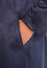 Marinblå mockajacka med en sidoficka och mjuk textur, med en närbild av en hand som vilar i fickan.