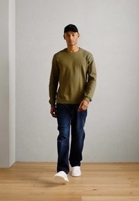Sweatshirt verde măsliniu, blugi albaștri, adidași albaștri, șapcă neagră; model mergând pe un podea din lemn, pe un fundal gri deschis simplu.