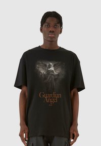 Mustan puuvillapaidan etupuolella on suuri kuva suojelusenkeli siipineen, ja sen alapuolella on oranssi teksti "Guardian Angel". Lyhyet hihat, rento istuvuus.