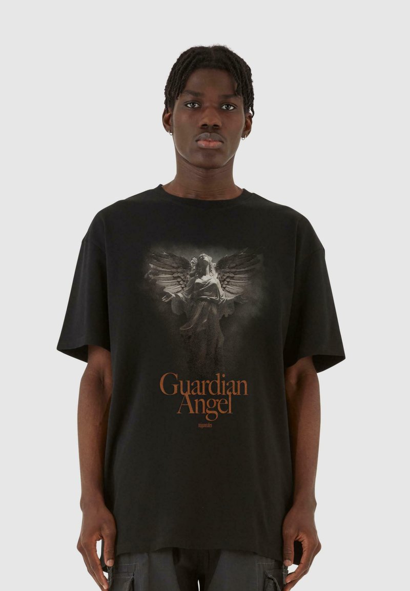 Mustan puuvillapaidan etupuolella on suuri kuva suojelusenkeli siipineen, ja sen alapuolella on oranssi teksti "Guardian Angel". Lyhyet hihat, rento istuvuus.