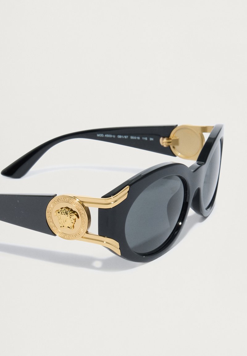 Schwarze Sonnenbrille mit glänzender Oberfläche, ausgestattet mit goldenen Akzenten und einem Medusa-Emblem an den Schläfen. Dunkle Gläser und runde Form.