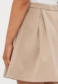 Falda plisada beige en tejido texturizado, con una cinturilla ajustada y forma fluida, con sutiles detalles de patrón visibles en el material.