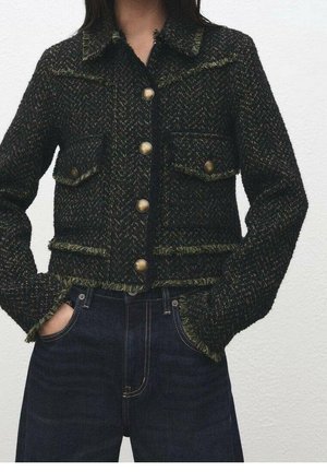 Femme portant une veste texturée vert foncé et noir à boutons en laiton et un jean bleu foncé, les mains dans les poches.