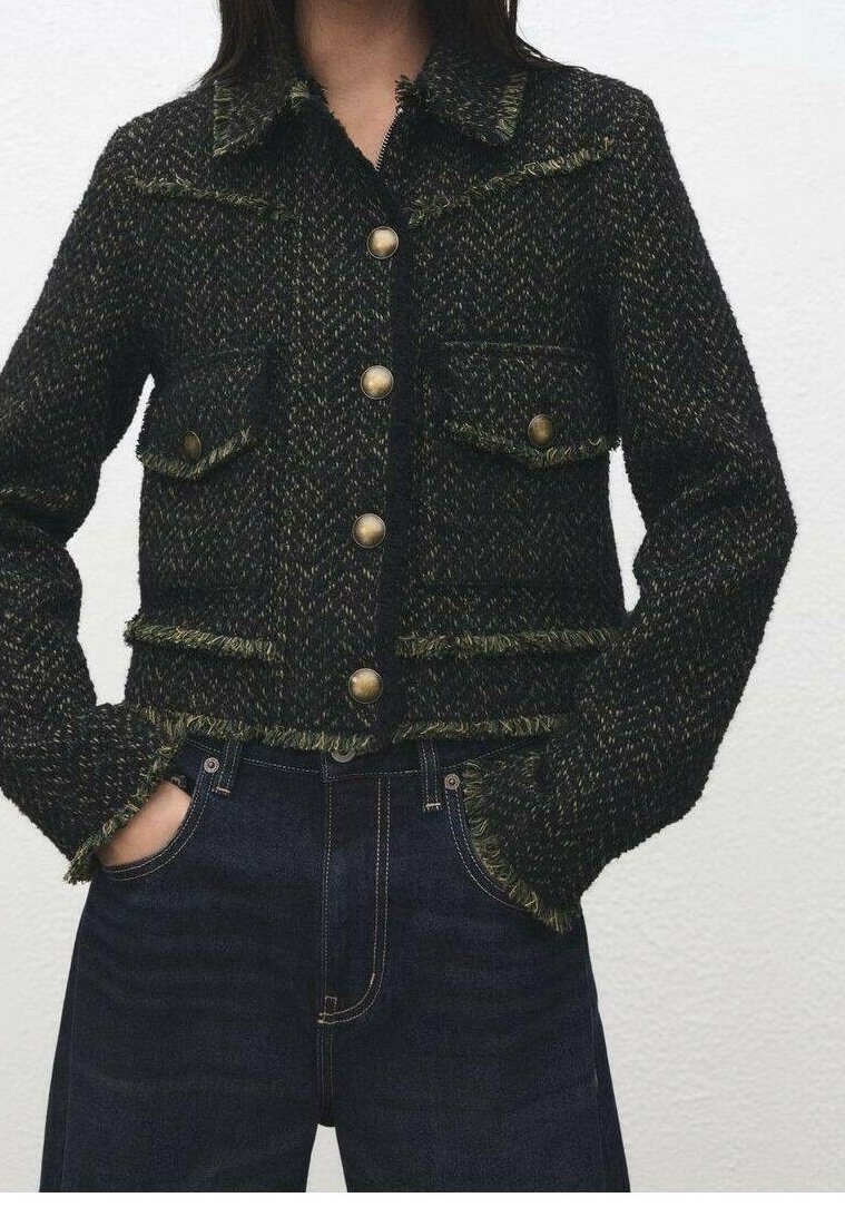 Femme portant une veste texturée vert foncé et noir à boutons en laiton et un jean bleu foncé, les mains dans les poches.