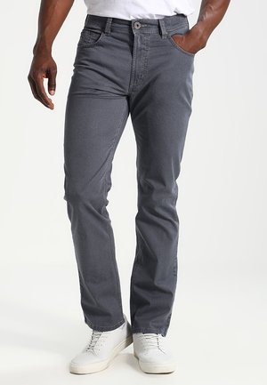 Man draagt nauwsluitende donkergrijze broek en witte sneakers, staat met één hand in de zak tegen een eenvoudige witte achtergrond.